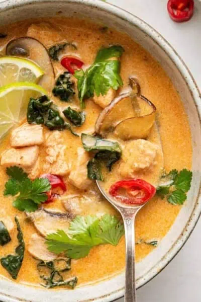 Close up of a spoon in a bowl of Thai coconut curry soup, garnished with chilis, lime slices, and cilantro