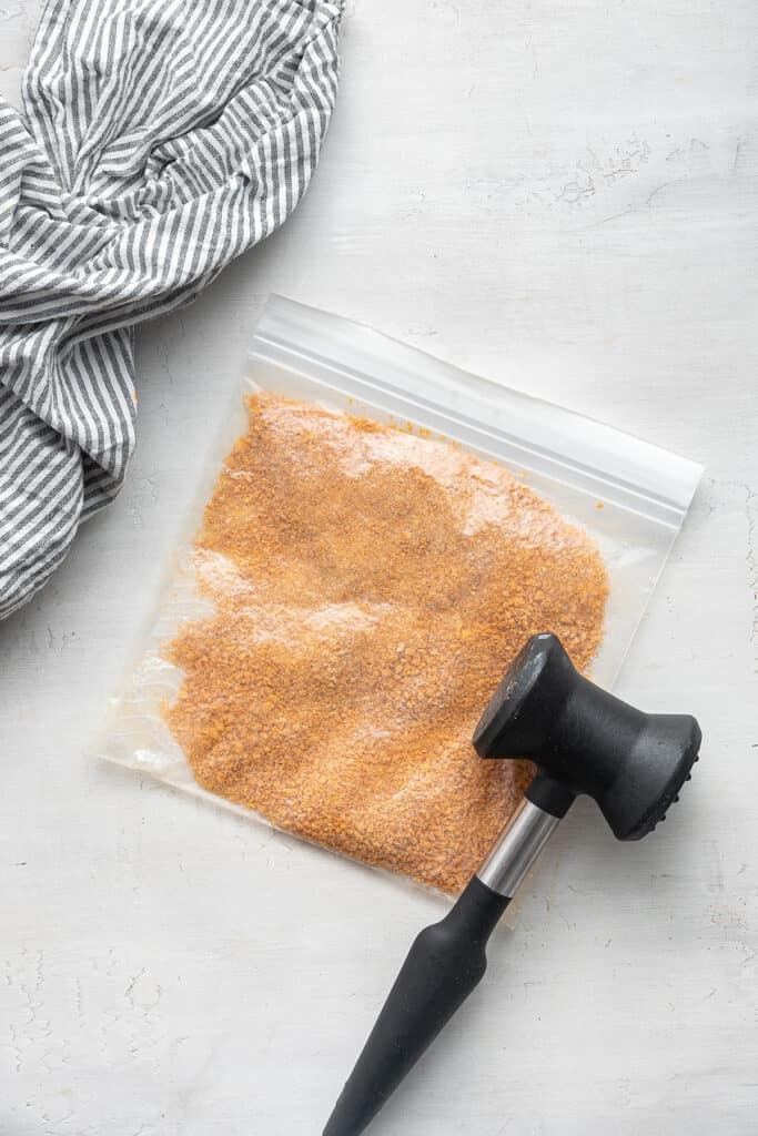 A ziplock bag filled with crushed corn flakes, with a meat tenderizer on top of it, next to a kitchen towel