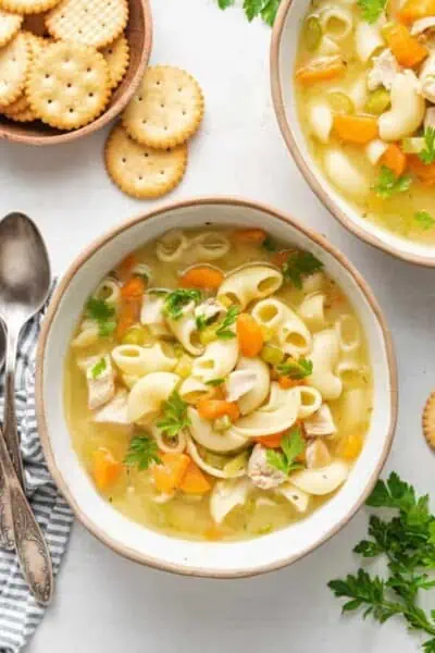 A bowl of chicken noodle soup garnished with parsley, next to another bowl, some round crackers, a napkin, and a spoon