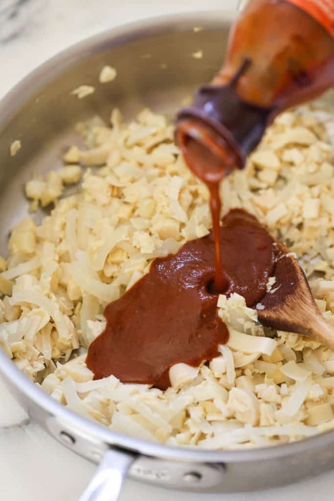 Pouring barbecue sauce into pan of hearts of palm and onion for vegan pulled pork