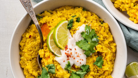 Overhead view of a bowl of kitchari topped with yogurt, cilantro, and lime slices, with a spoon in it, next to another bowl.