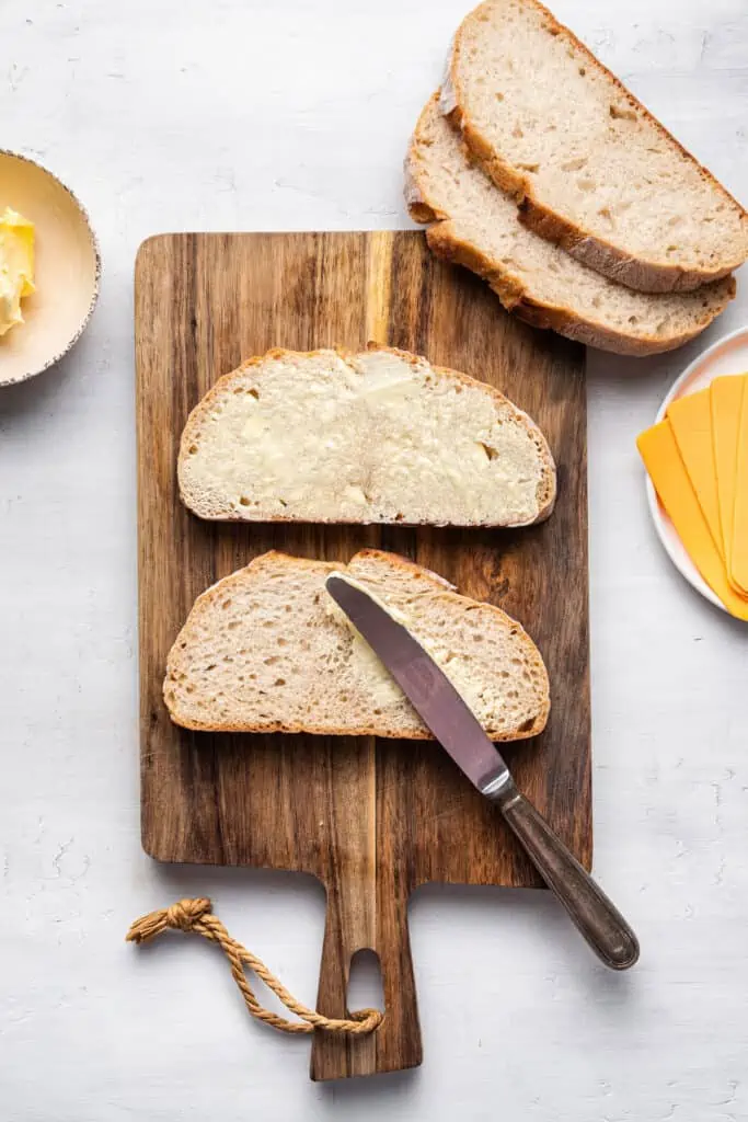 Butter is spread over one side of each bread slice on a wooden cutting board.