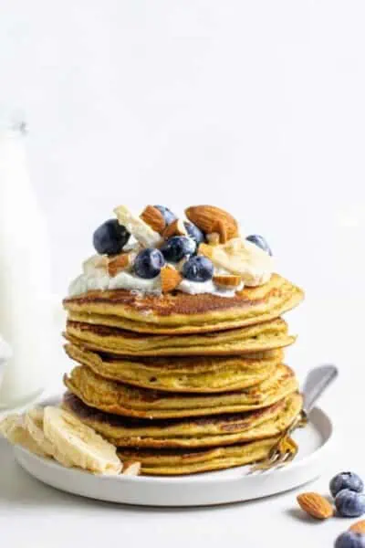 A stack of protein pancakes on a plate topped with fresh fruit and nuts.