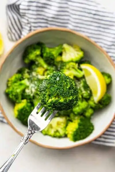 A fork holds a broccoli floret over a bowl of steamed broccoli garnished with nutritional yeast and a lemon wedge.