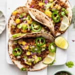 Three vegan tacos on white board with garnishes and lime wedges