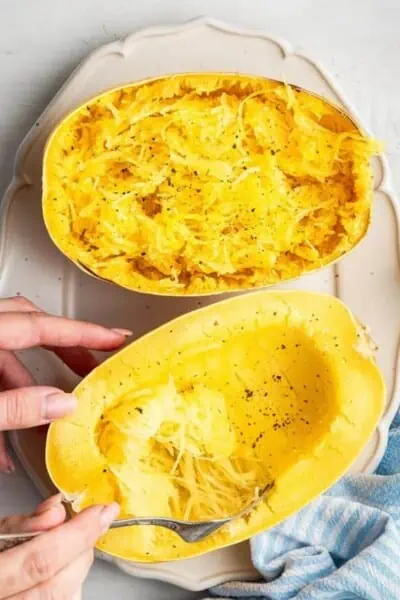 A cooked spaghetti squash cut in half on a serving tray, with one half shredded, and the other half in the process of being shredded by a fork