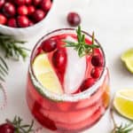 Overhead view of a cranberry margarita in a salt-rimmed glass, topped with a lime wedge, cranberries, and a sprig of rosemary, next to a bowl of cranberries and some rosemary sprigs