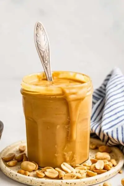 A jar of honey roasted peanut butter on a plate, with a spoon sticking out of it, peanuts on the plate, and a blue and white striped kitchen towel