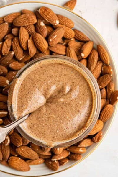 Overhead view of a jar of pumpkin spice almond butter with a spoon submerged in it, sitting on a plate covered in almonds