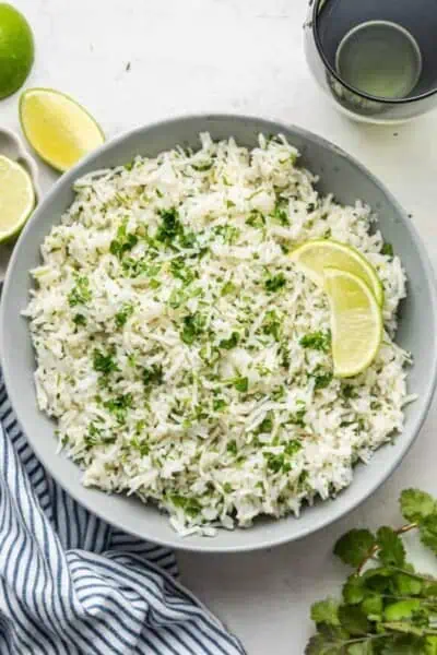 A plate of cilantro lime rice topped with lime slices, next to a sprig of cilantro, fresh limes, a kitchen towel, and a water glass