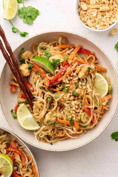 Overhead view of a bowl of chicken pad tai with lime wedges and chopsticks on top, next to a bowl of peanuts, and some lime slices and cilantro