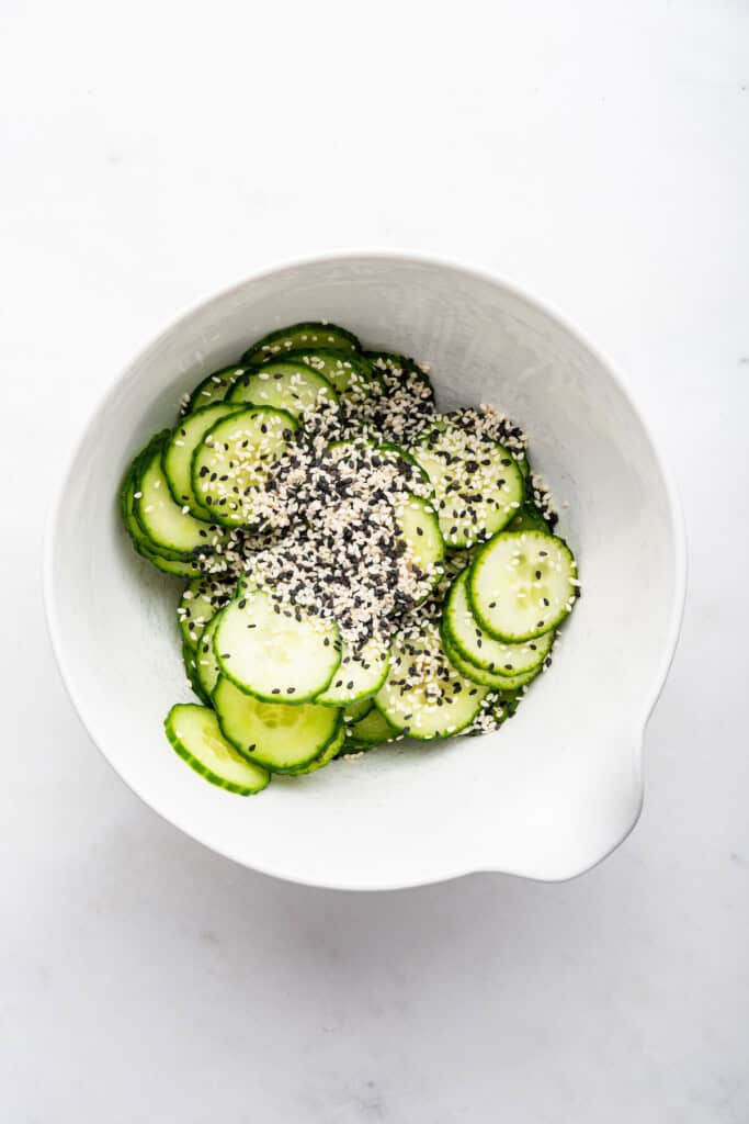 Overhead view of sesame seeds added to bowl of cucumbers