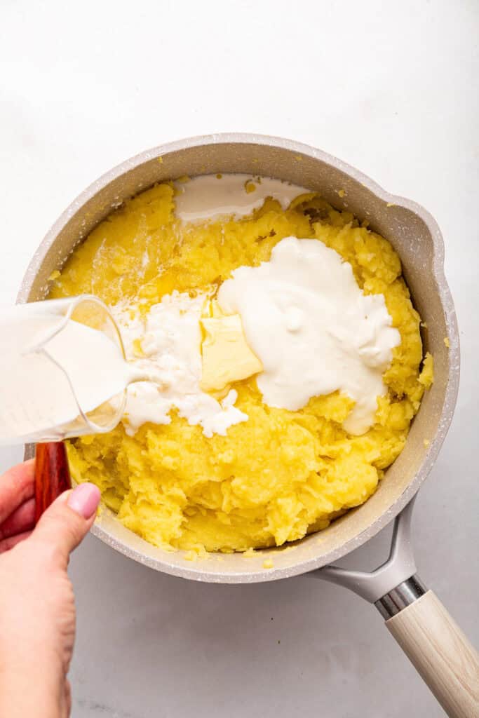 Adding vegan milk, butter, and yogurt to dairy-free mashed potatoes in pan