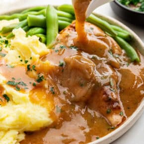 Pouring gravy over smothered chicken on plate with mashed potatoes and green beans