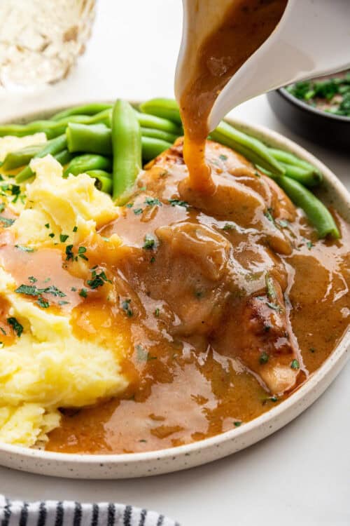 Pouring gravy over smothered chicken on plate with mashed potatoes and green beans